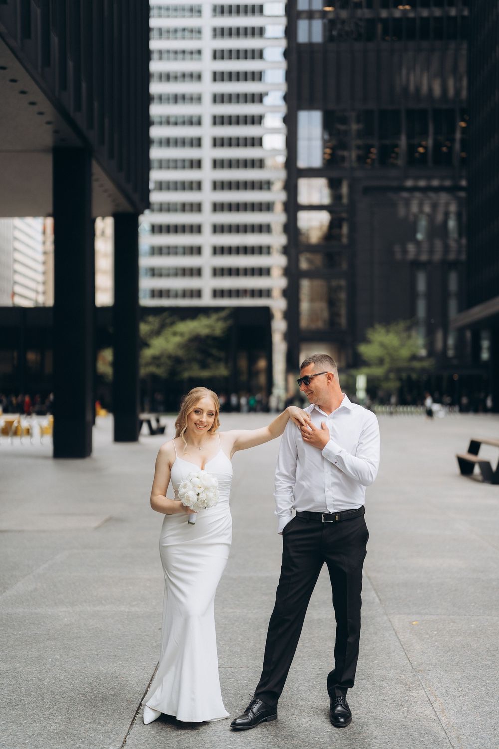 A couple in wedding attire dance together on a city plaza with modern office buildings in the background.