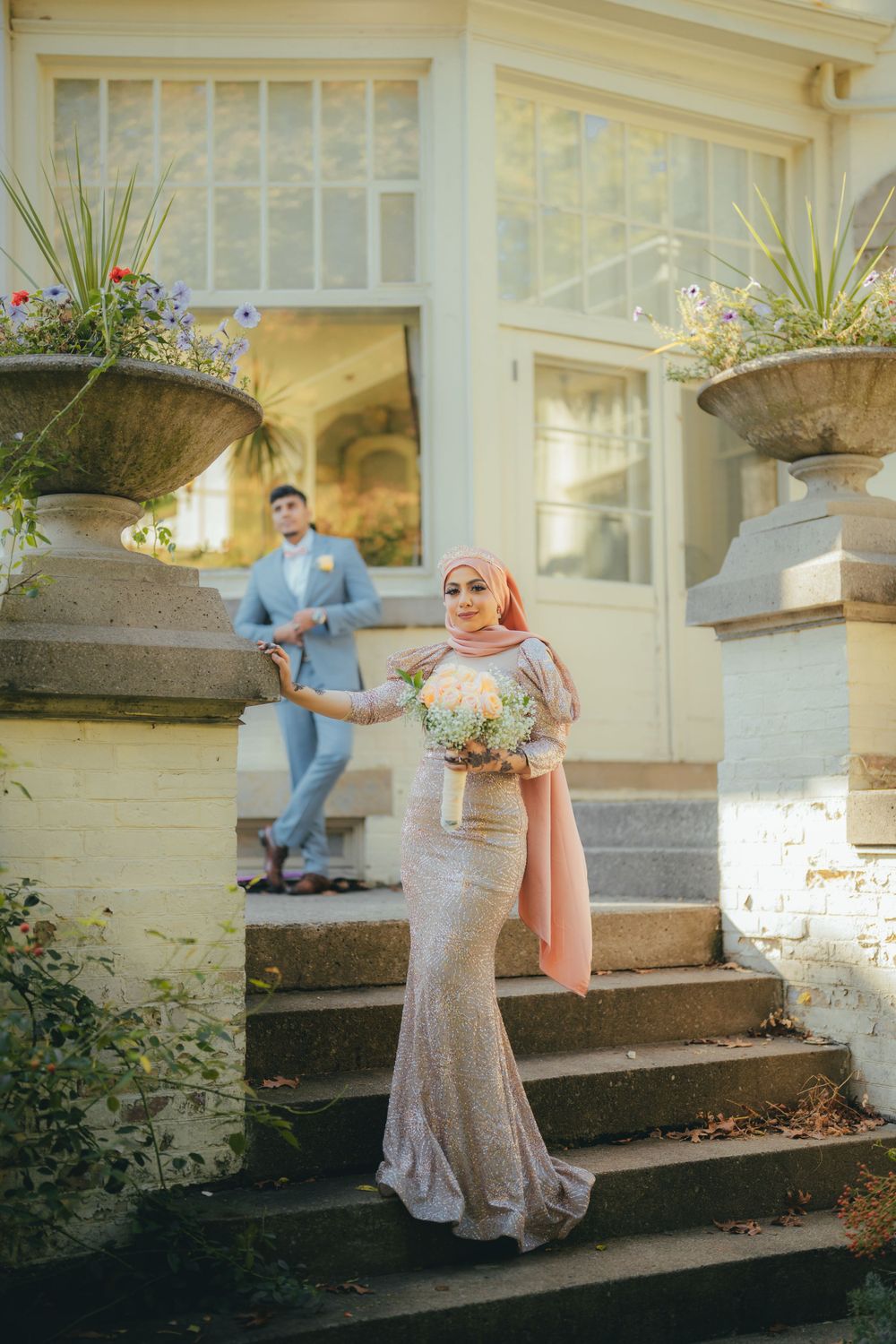 A couple in formal attire pose on elegant stone steps with decorative planters and flowers outside a building in sunlight.