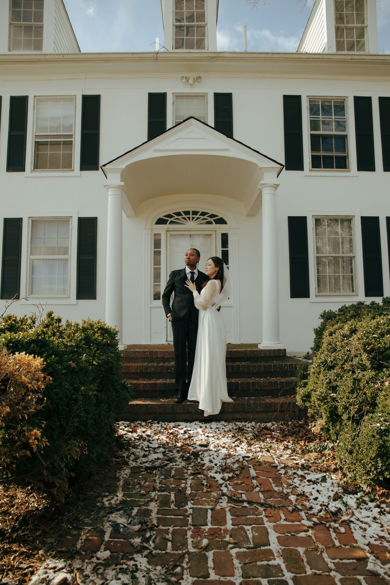 Elegant wedding couple stands on brick walkway in front of stately white colonial house with black shutters and portico.