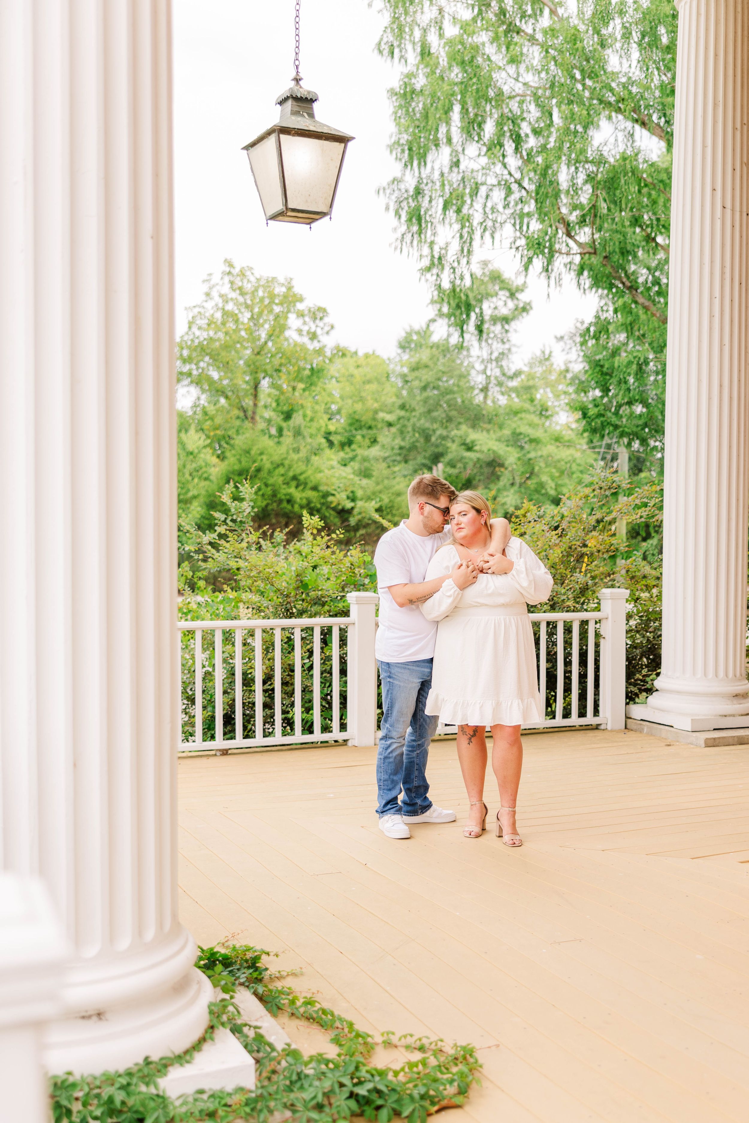 Couple poses between elegant white columns on outdoor veranda with greenery.