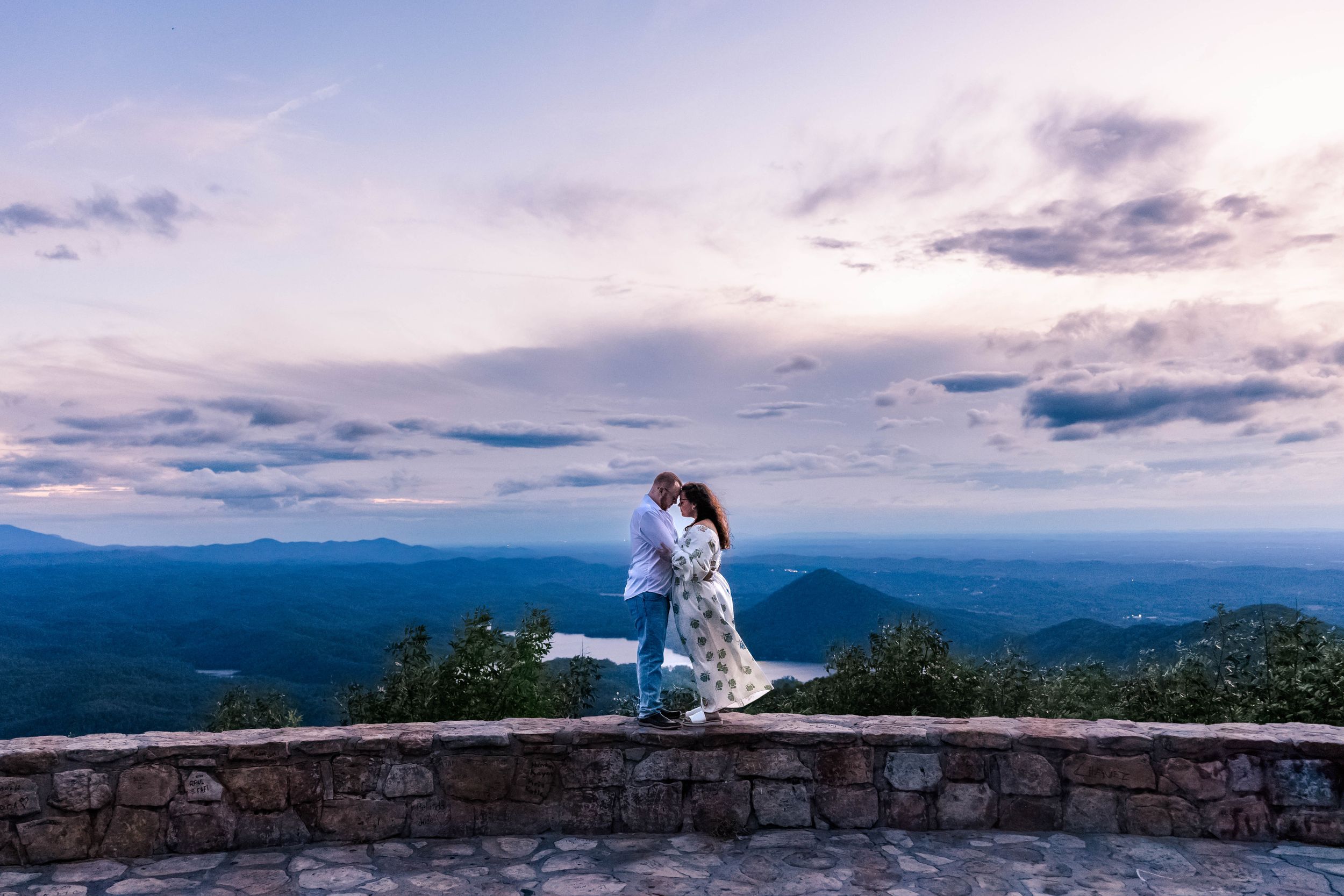 Couple sharing a romantic moment on stone wall overlooking scenic mountain landscape at dusk.