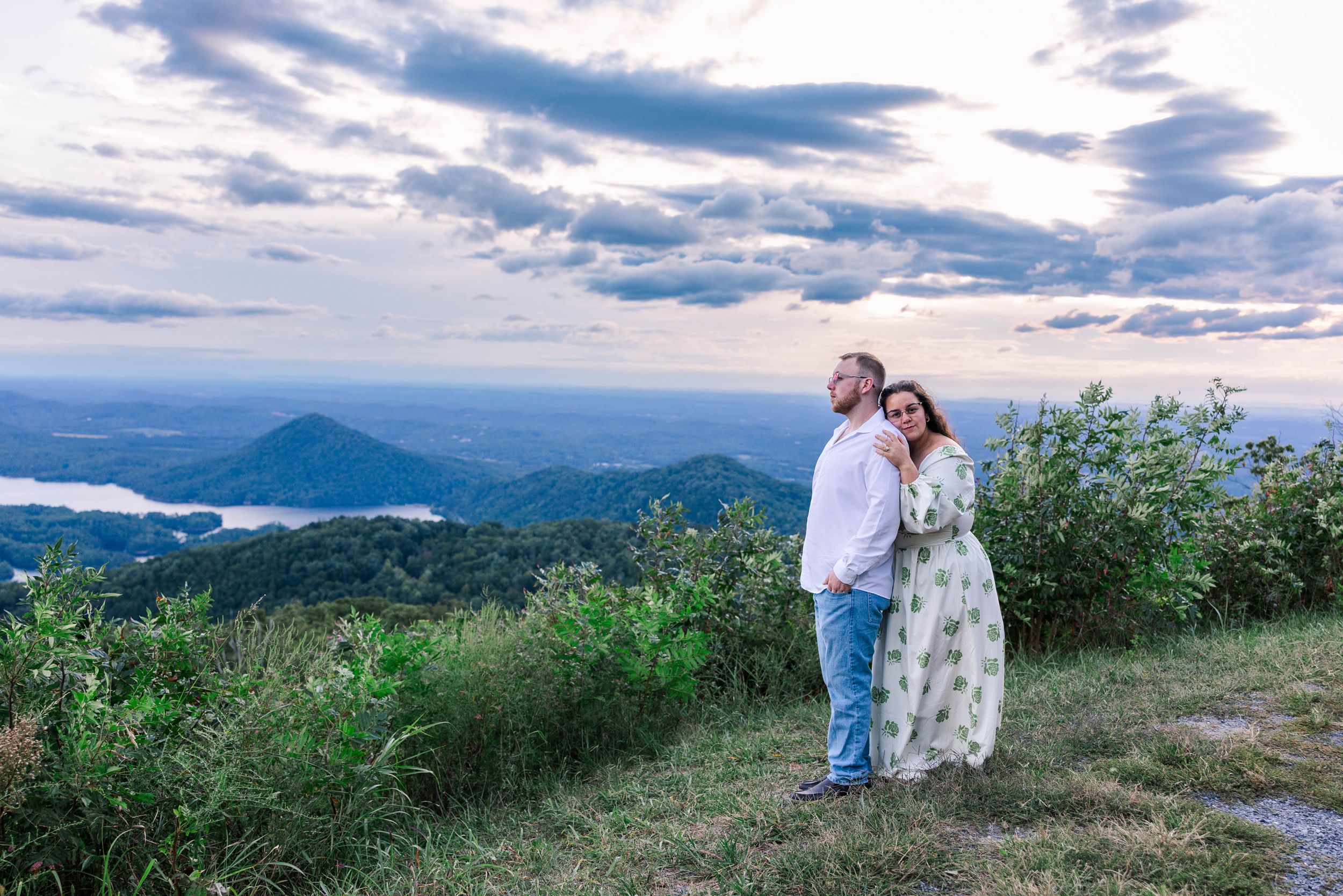 A couple stands together on a mountain overlook with scenic rolling hills and dramatic clouds in the background.