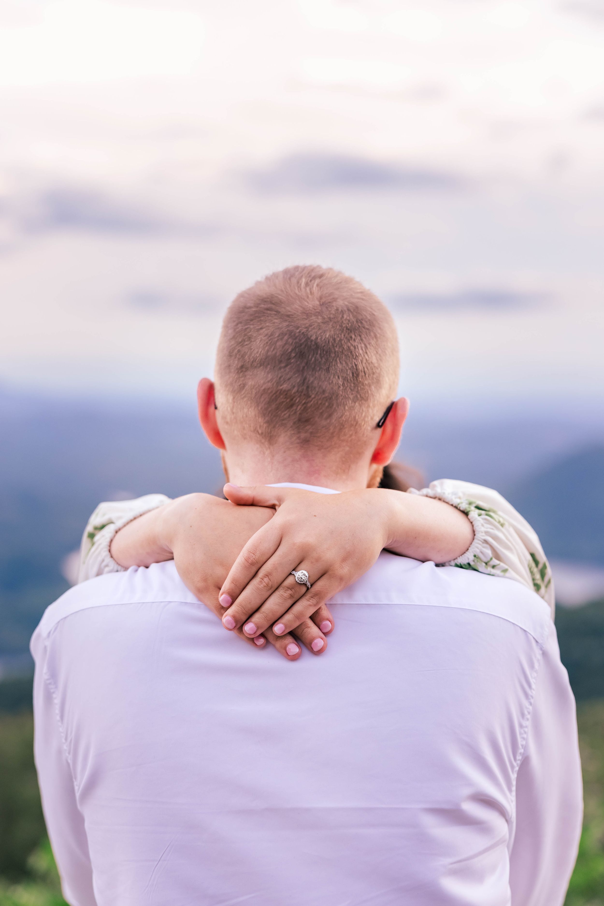 Intimate back view with arms wrapped around showing off an engagement ring against mountain backdrop.