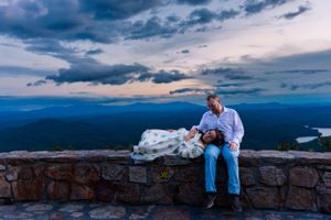 A couple sits together on a stone wall at sunset with dramatic mountain vistas stretching into the distance.