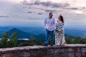 Couple standing on stone wall overlook with sweeping mountain views and cloudy sky backdrop.