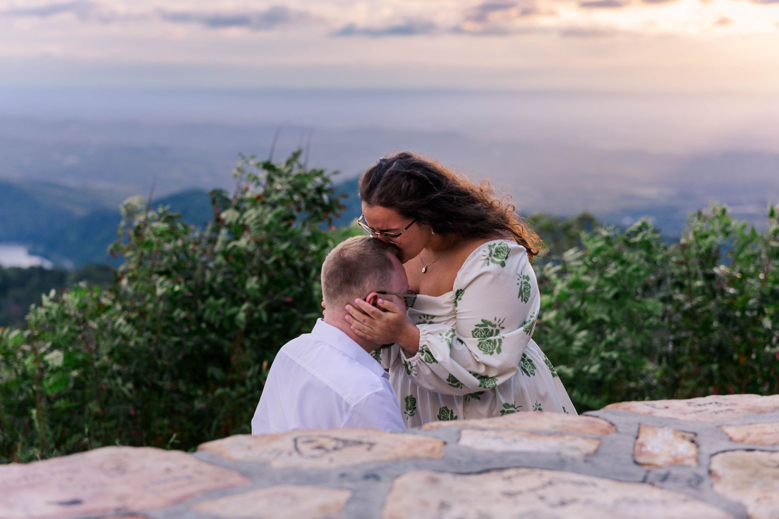 Intimate moment shared on mountain overlook with dramatic sunset sky and valley views.