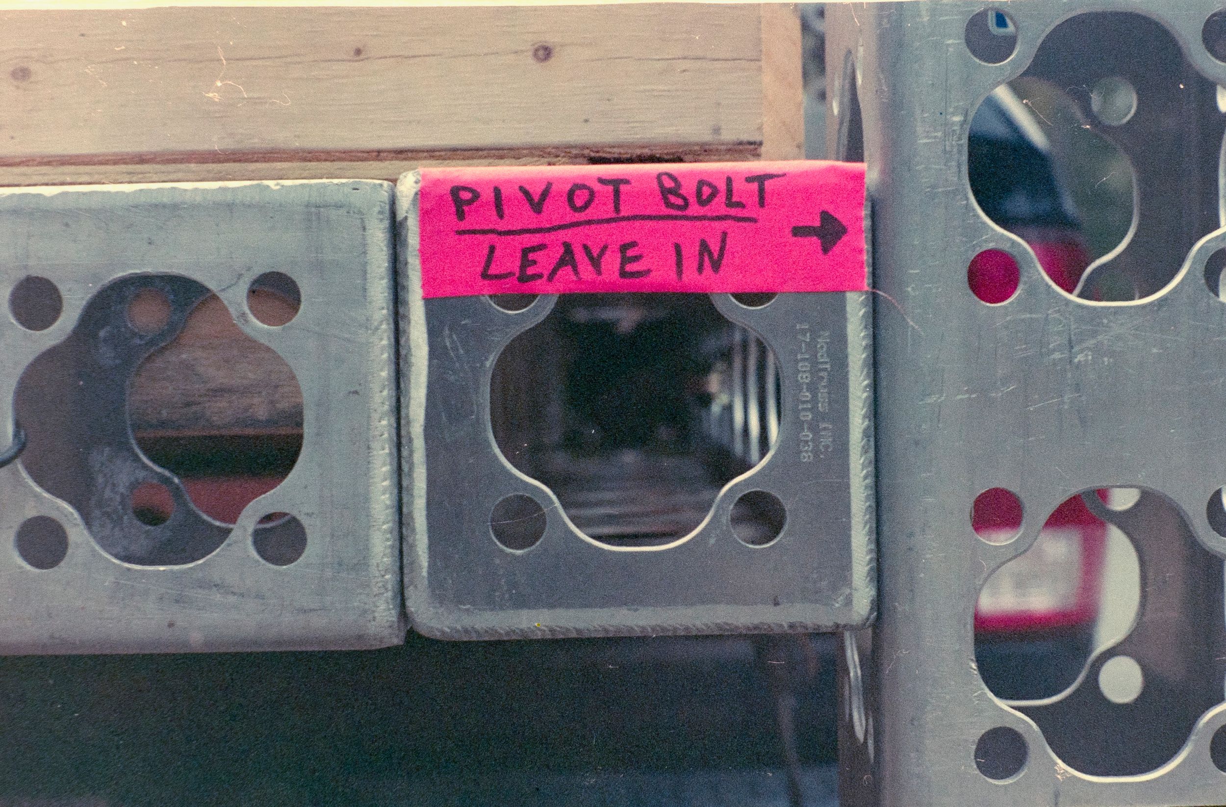 Close-up of metal truss with pink label reading 'Pivot bolt leave in'.