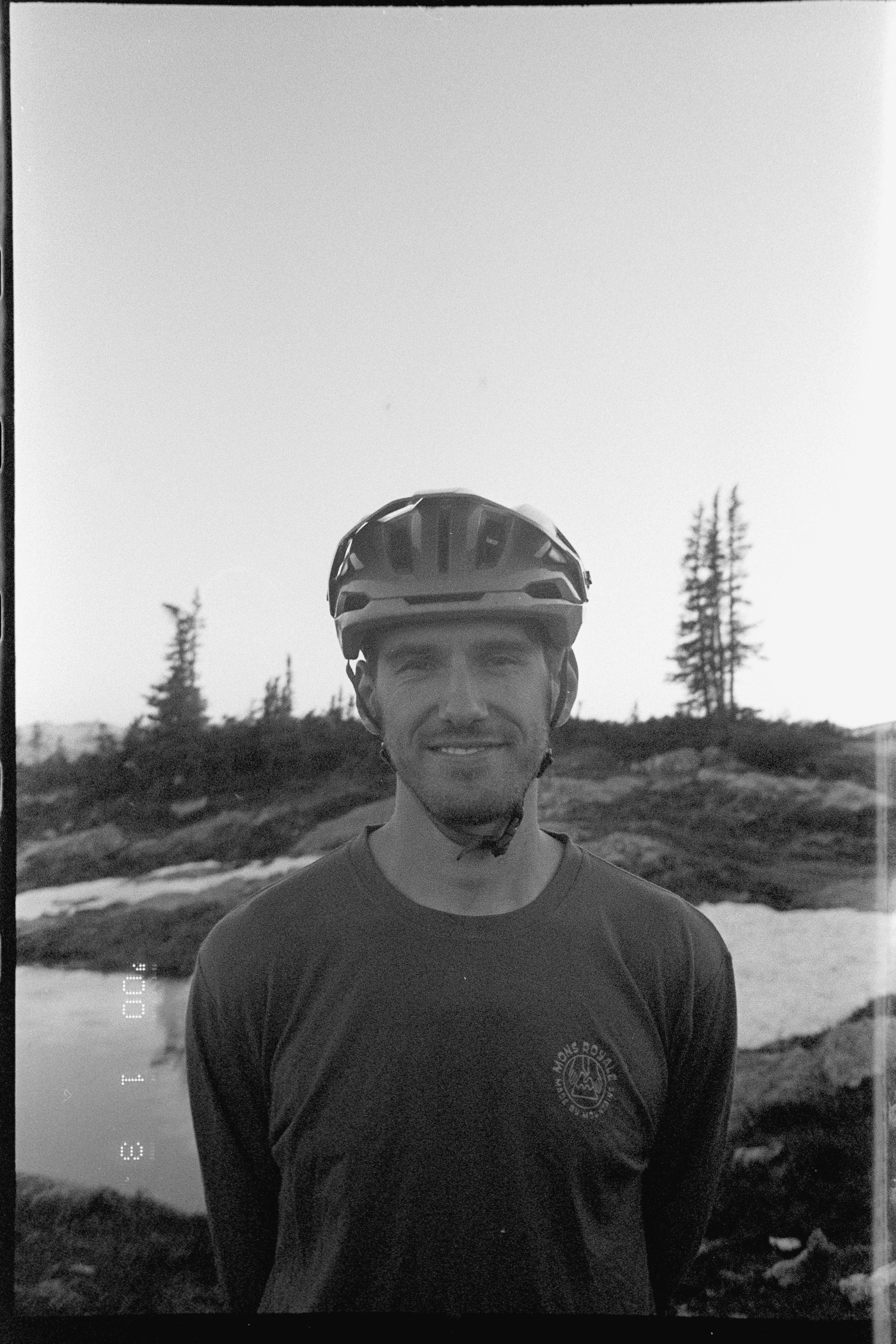 Black and white portrait of cyclist Joel Ducrot wearing a helmet with evergreen trees in the background.