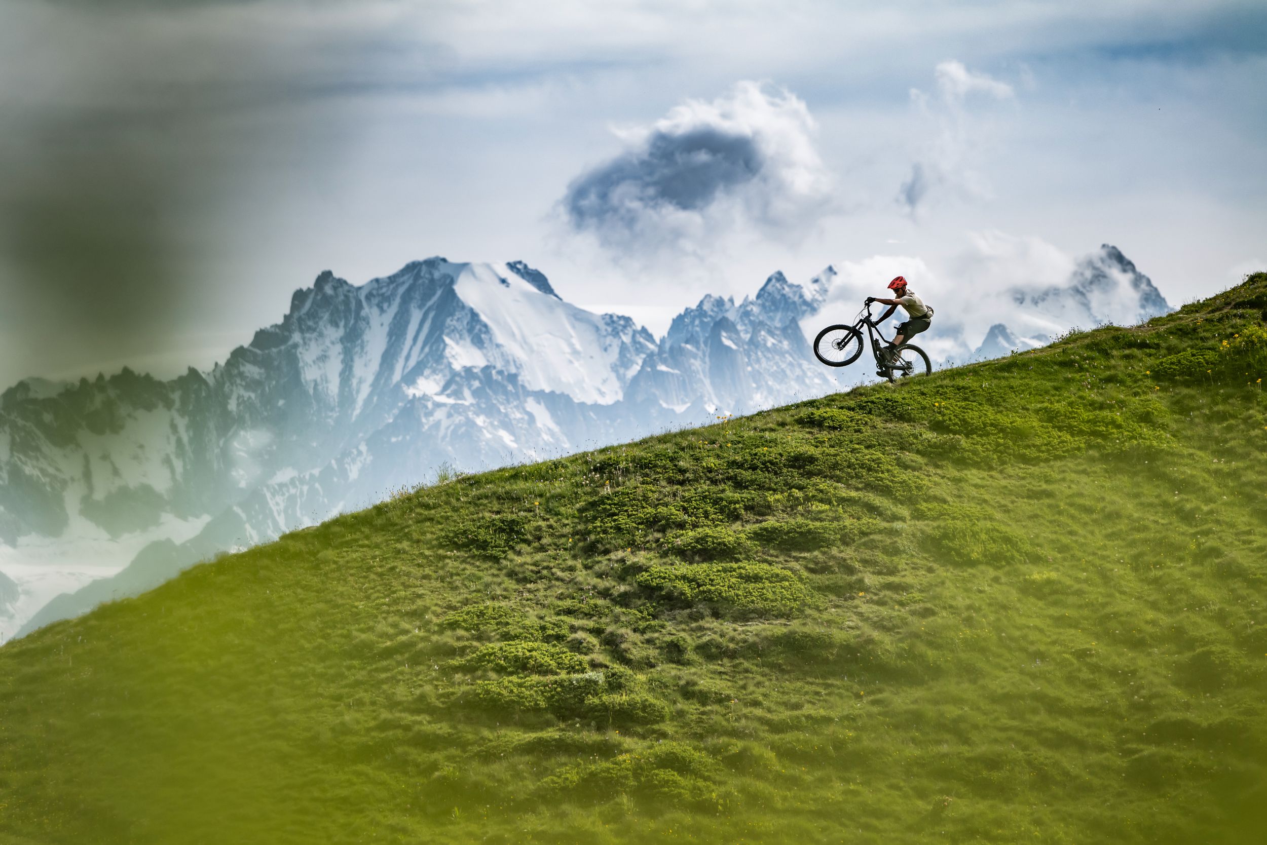 Mountain biker Joel Ducrot silhouetted against dramatic alpine peaks while riding across a grassy slope. Shot by Gaudenz Danuser