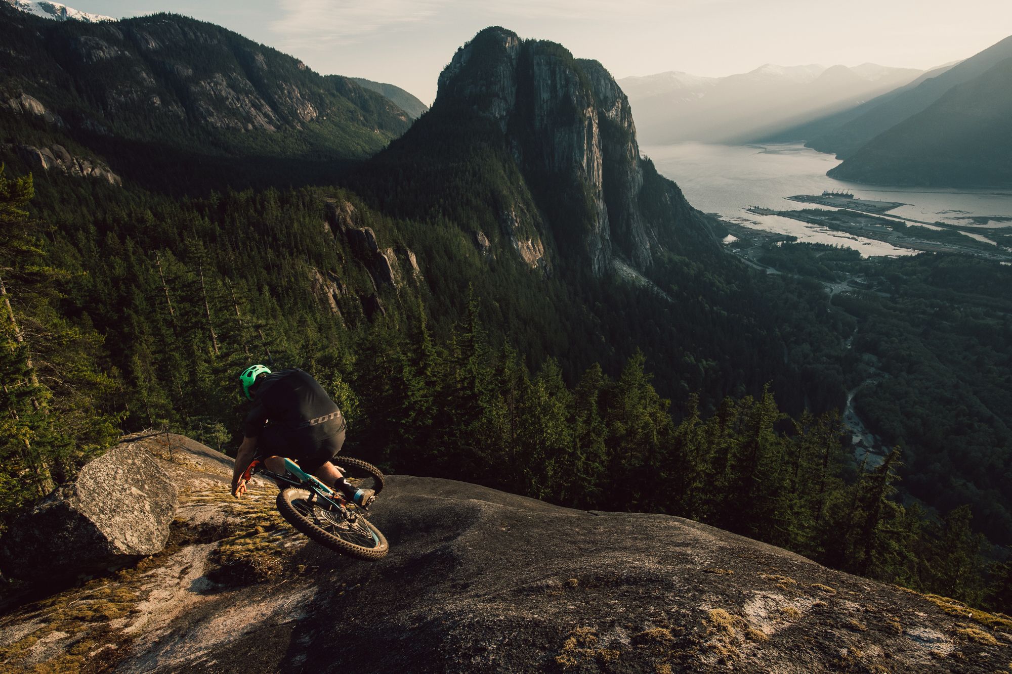 Mountain biker Joel Ducrot overlooking dramatic valley view witht Stawamus Chief from rocky outcrop during sunset.