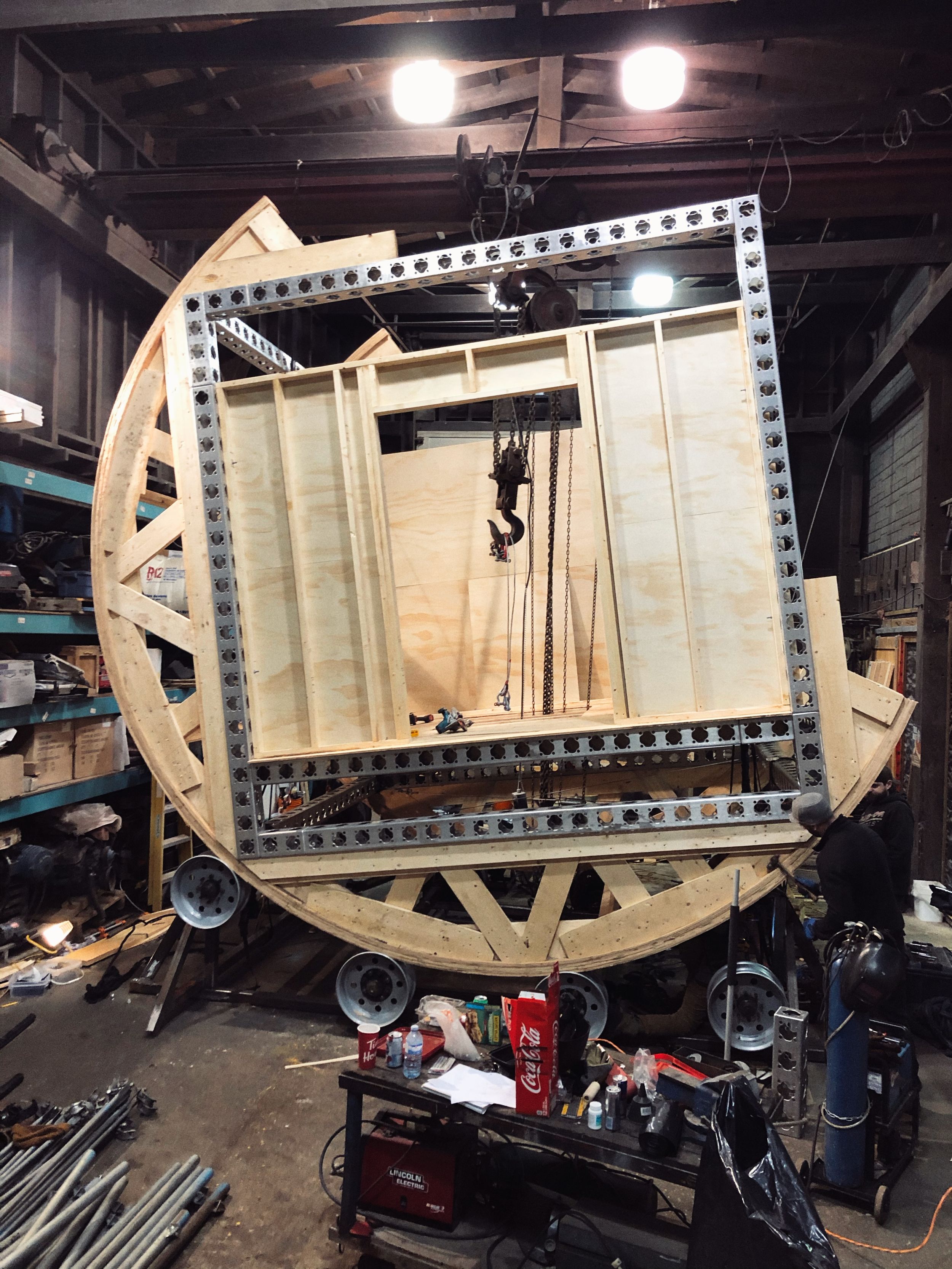 Large circular wooden frame structure to perform rotating room stunt in industrial workshop setting.