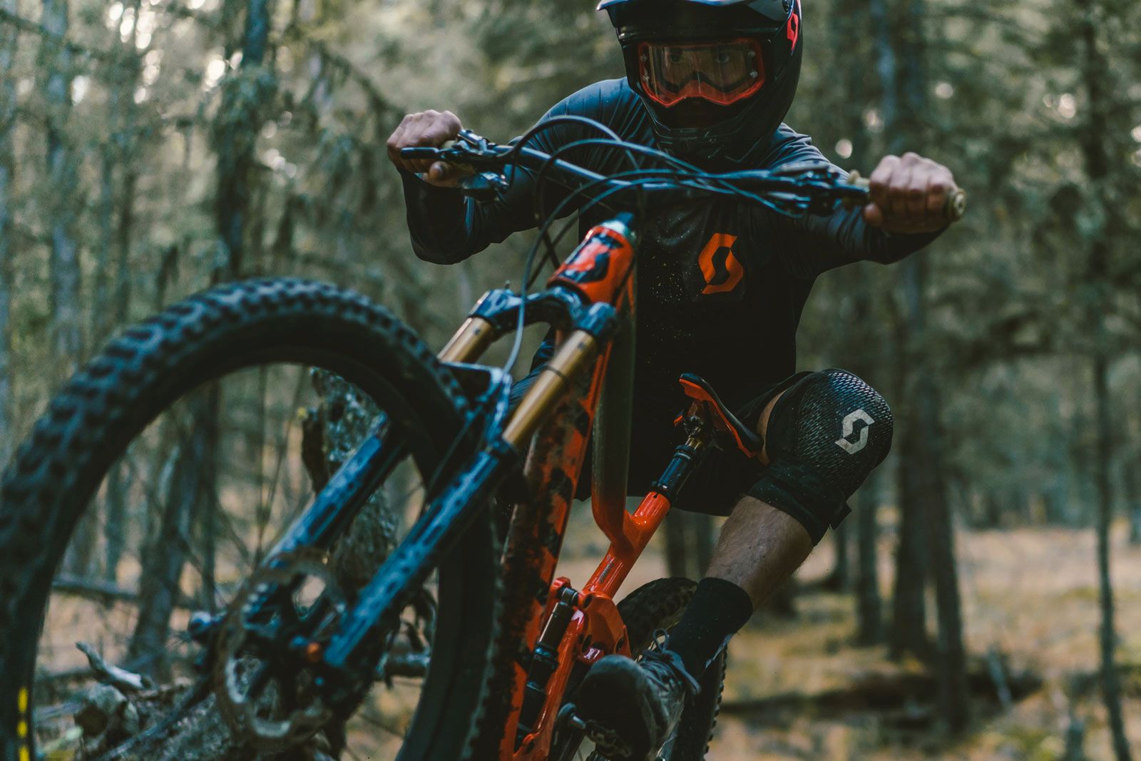 Mountain biker Joel Ducrot in full protective gear rides an orange and blue bike through a forest trail. Shot by Steve Shannon