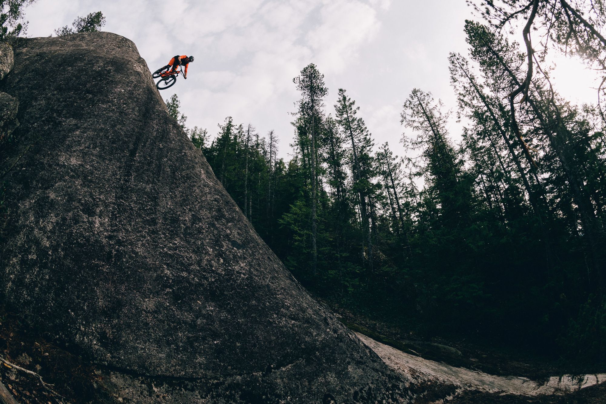 Mountain biker Joel Ducrot soaring off a steep rock jump against a forested backdrop. Shot by Margus Riga