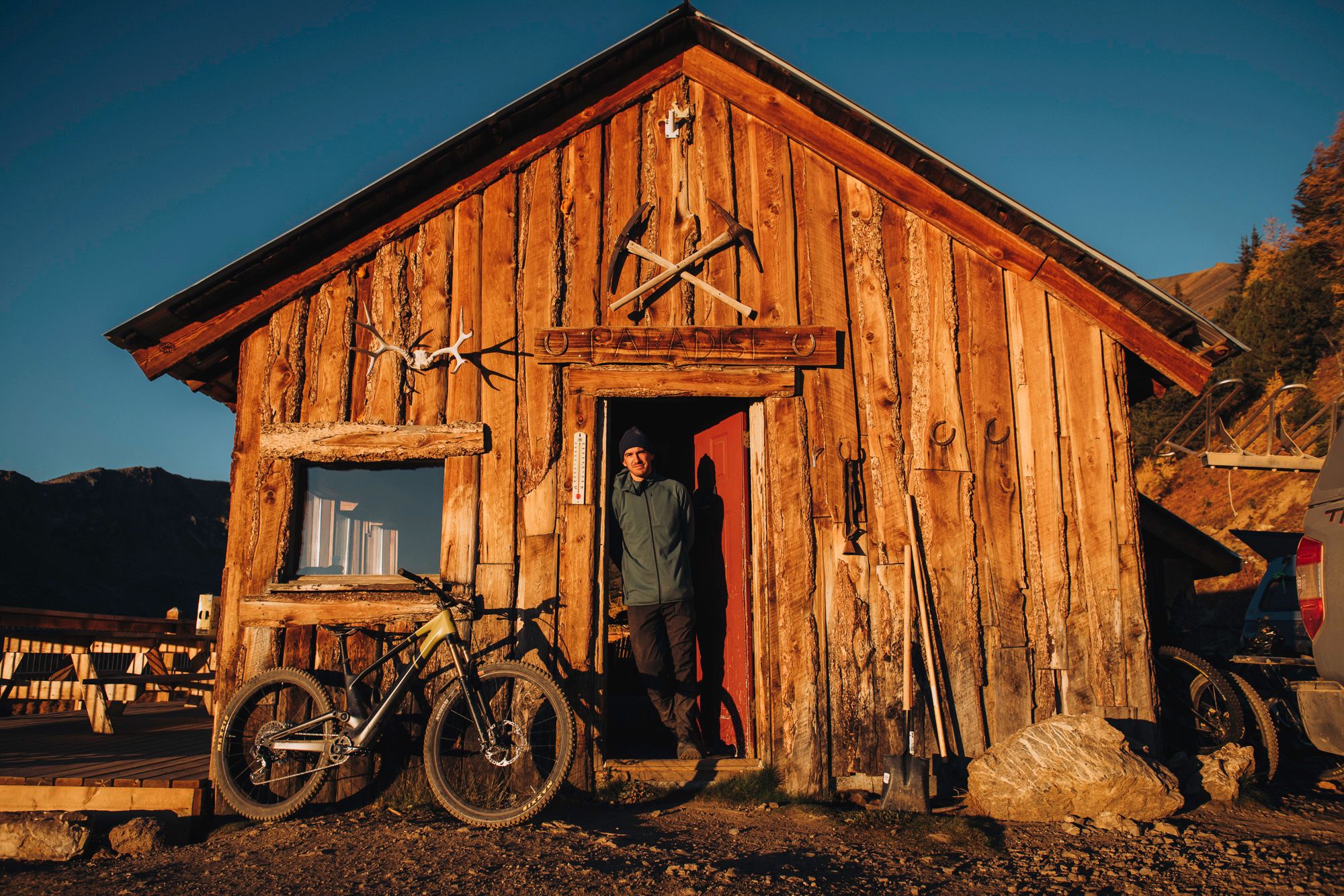 Rustic wooden cabin with mountain bike parked outside against mountain backdrop at sunset.