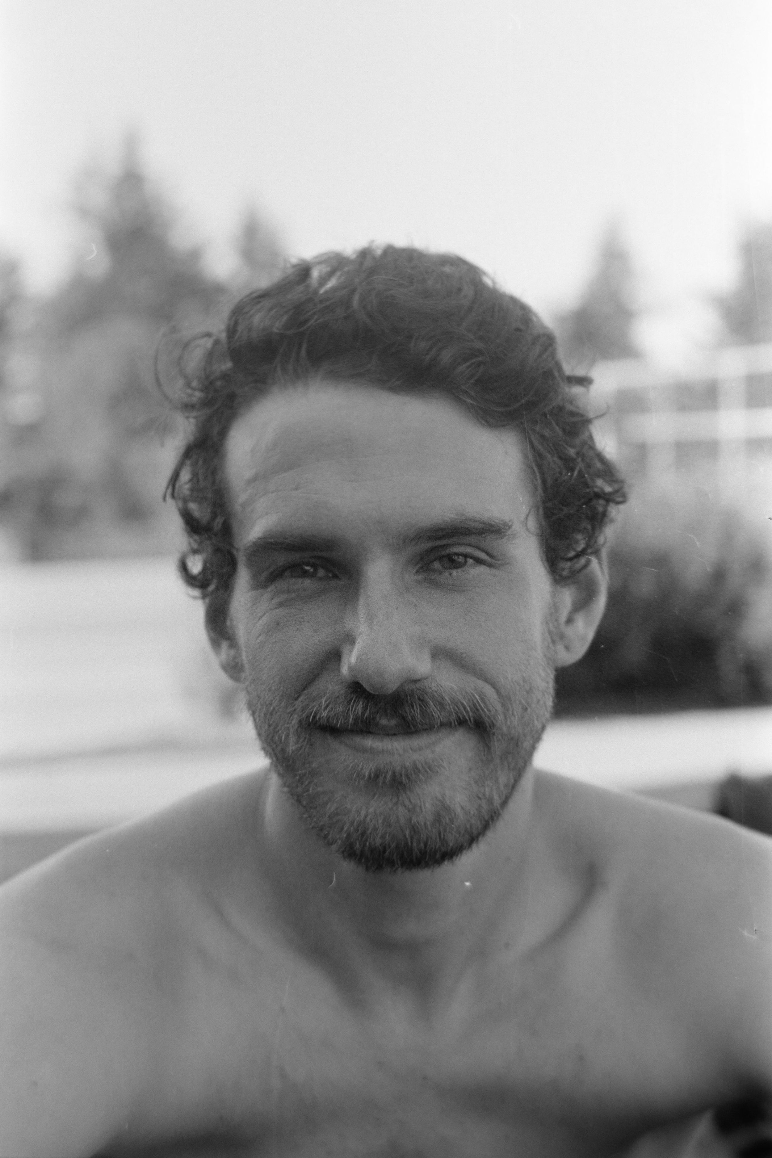 Black and white portrait of Joel Ducrot with curly hair and beard smiling at the camera.