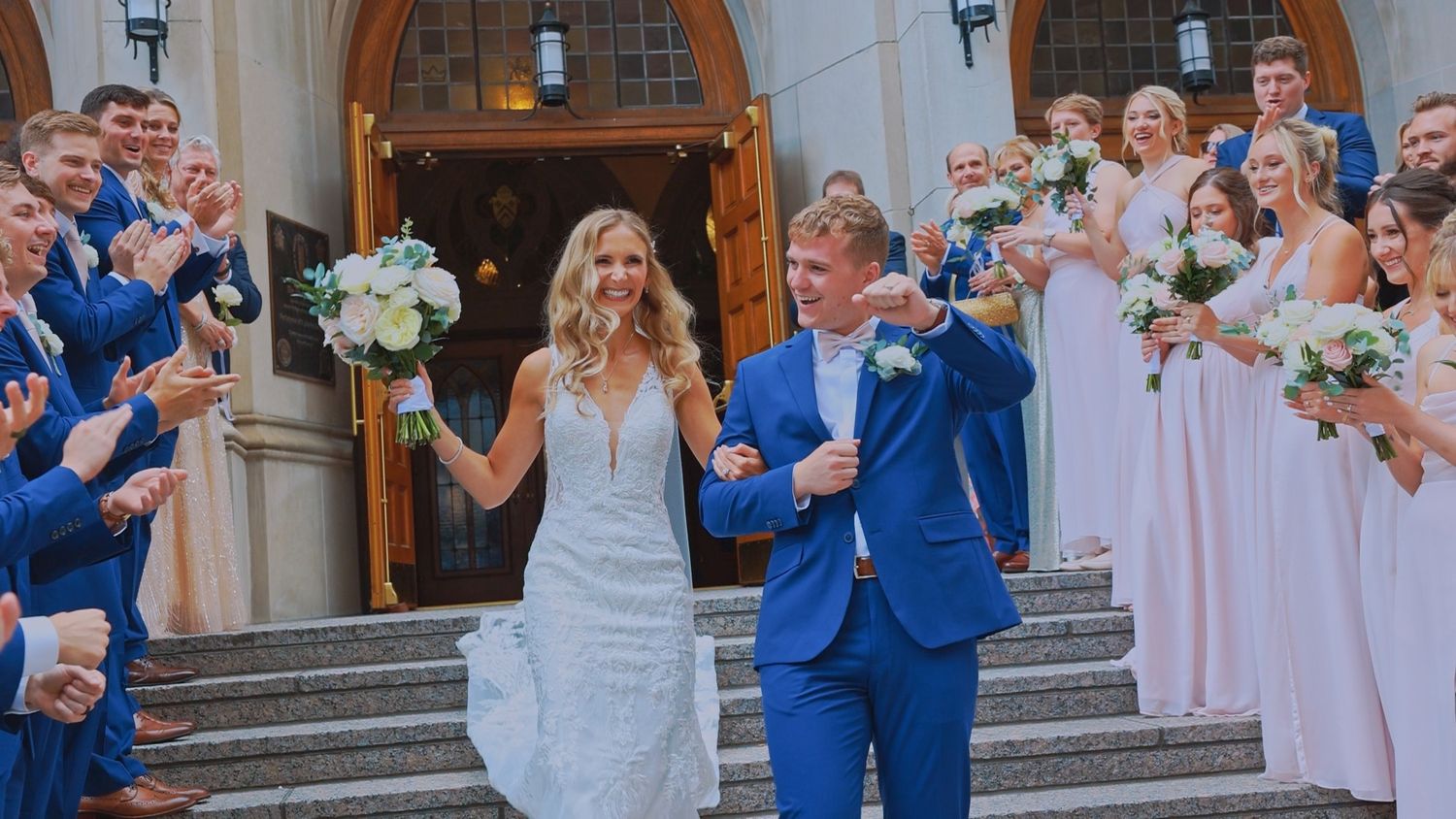 A joyful wedding party celebrates on church steps as newlyweds walk down in a white dress and bright blue suit.