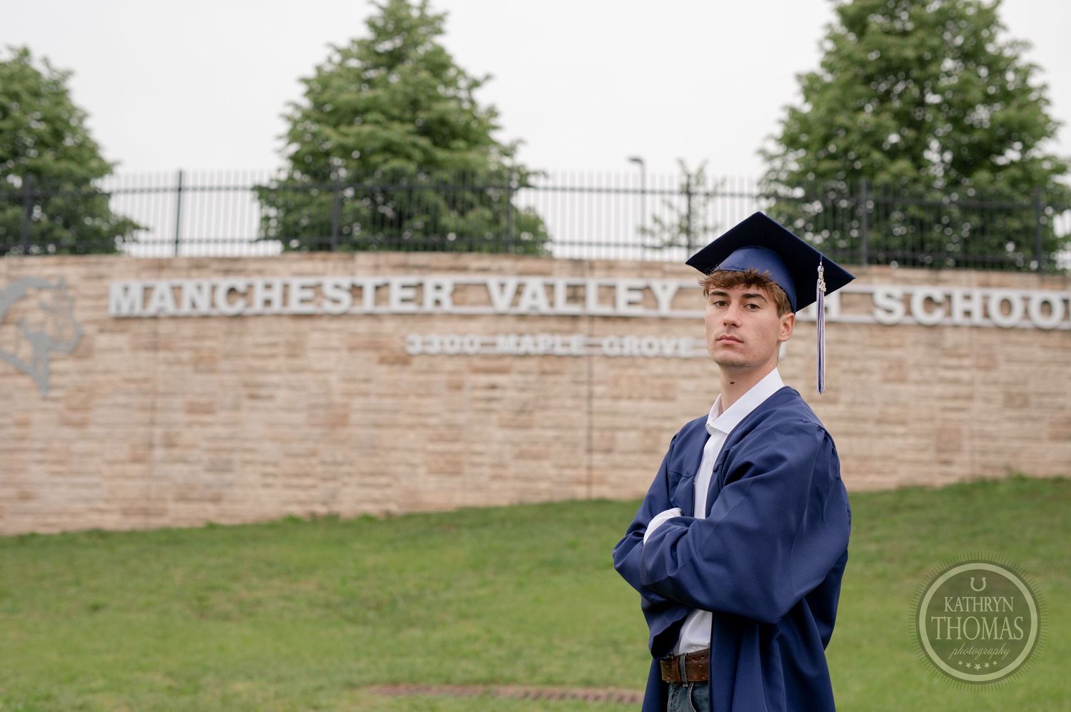 Graduate in traditional academic regalia poses in front of Manchester Valley School sign.