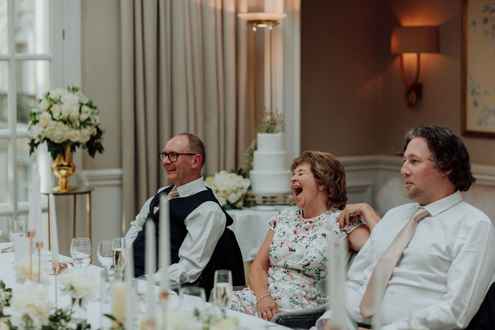 Wedding reception guests sharing a candid laughing moment at an elegant dinner table with white floral arrangements.