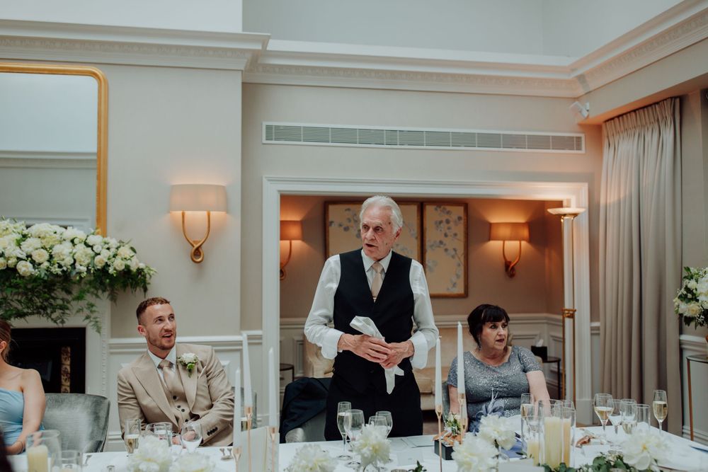 People seated at an elegant wedding reception dinner table while someone stands to deliver a speech.