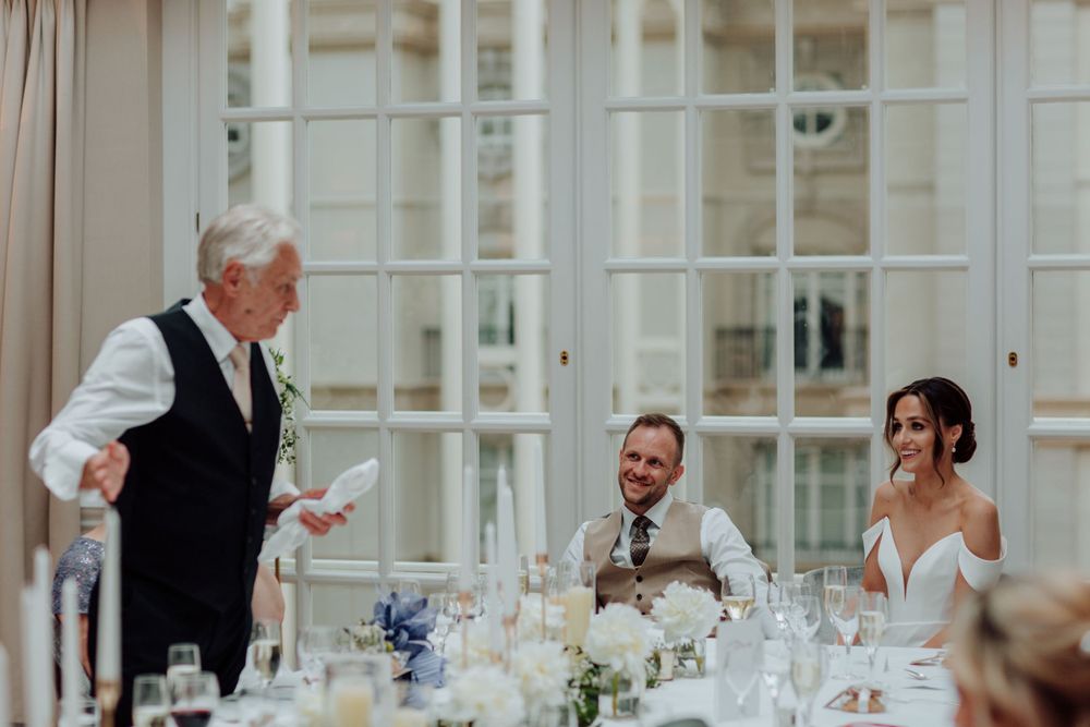 A joyful moment during a wedding reception as guests laugh during a dinner speech at an elegant venue with white decor.