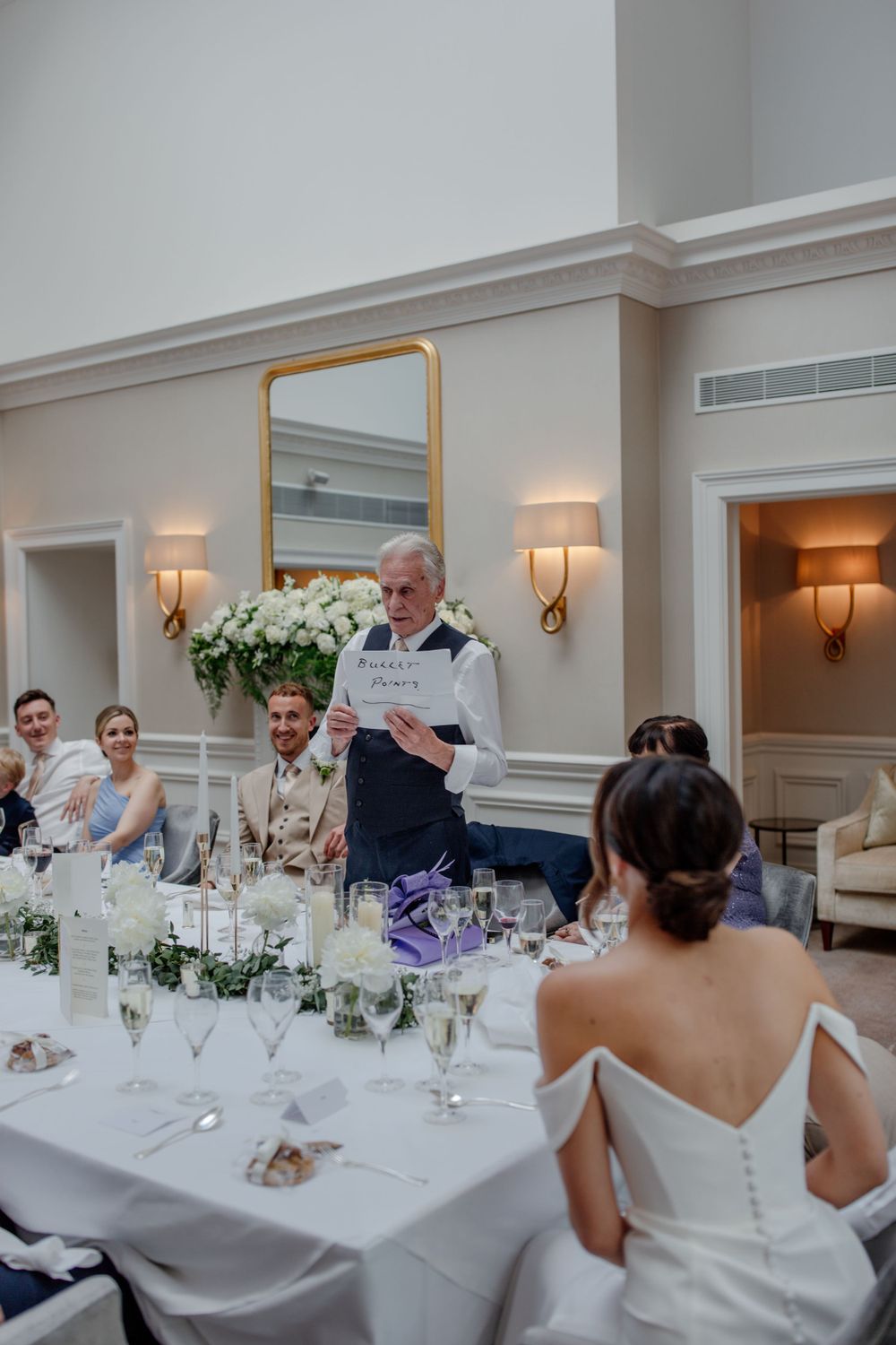 Wedding reception speech being given at an elegant white-tablecloth dining setting with floral centerpieces.