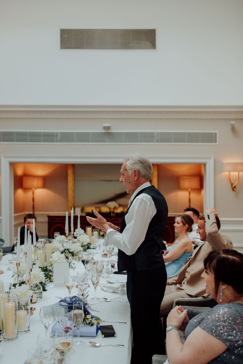 Speaker delivers a speech at an elegant wedding reception with white table settings and warm lighting.