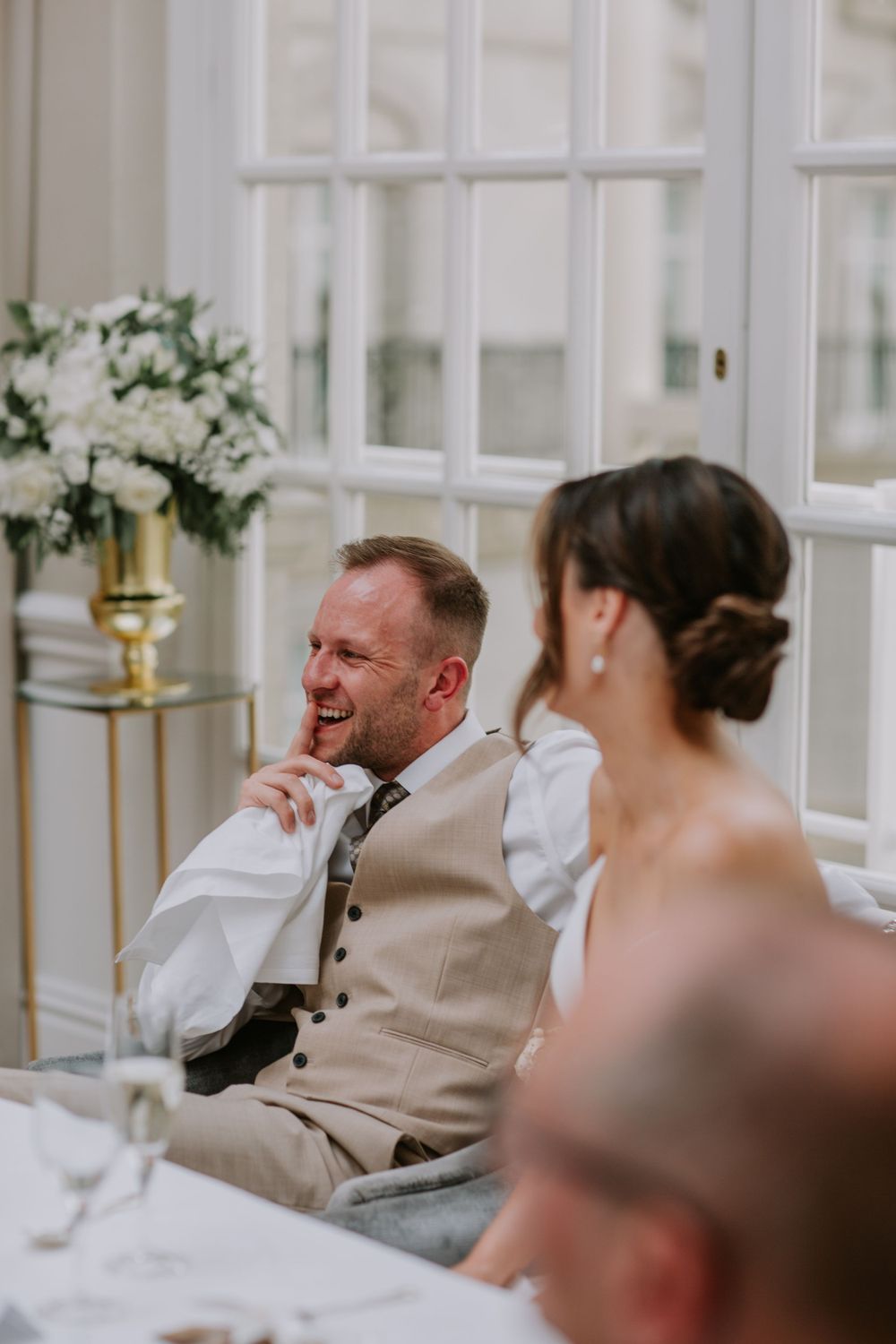 Wedding guests seated at reception with beige suit and laughing amid elegant decor with white floral arrangements.