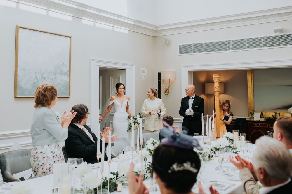 Wedding guests raise glasses during an elegant toast at a formal reception in a white-walled dining room.