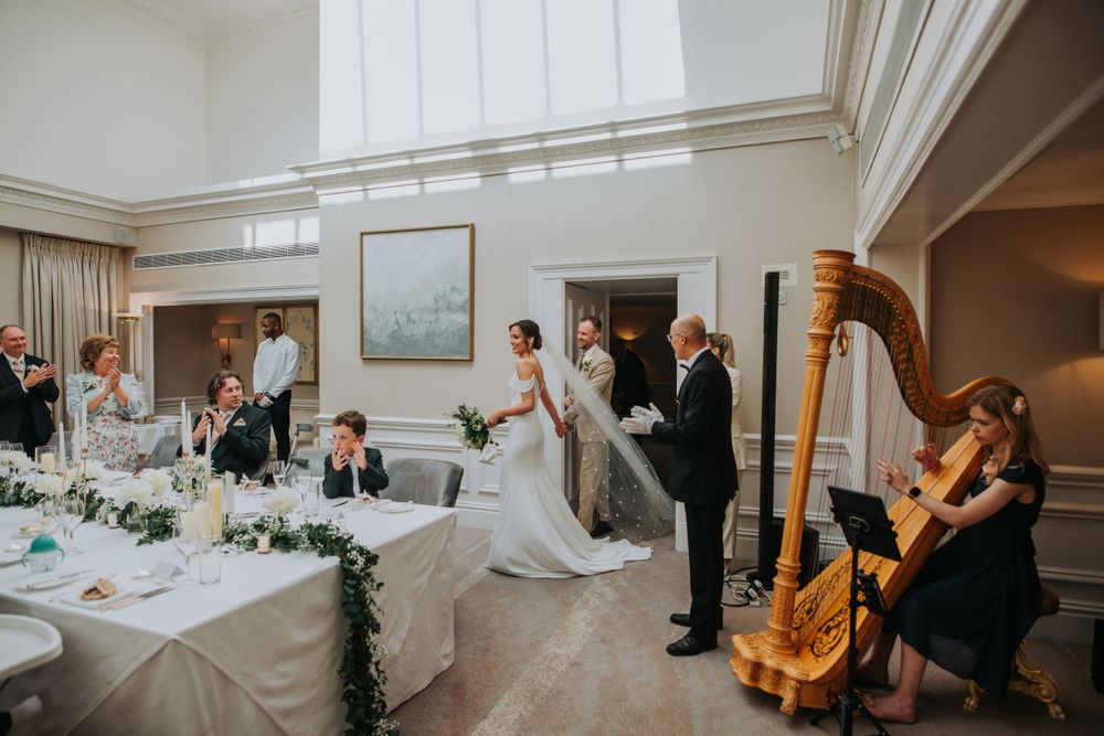 Elegant wedding reception scene in a bright room with white decor, long dining tables, and a harpist performing live music.