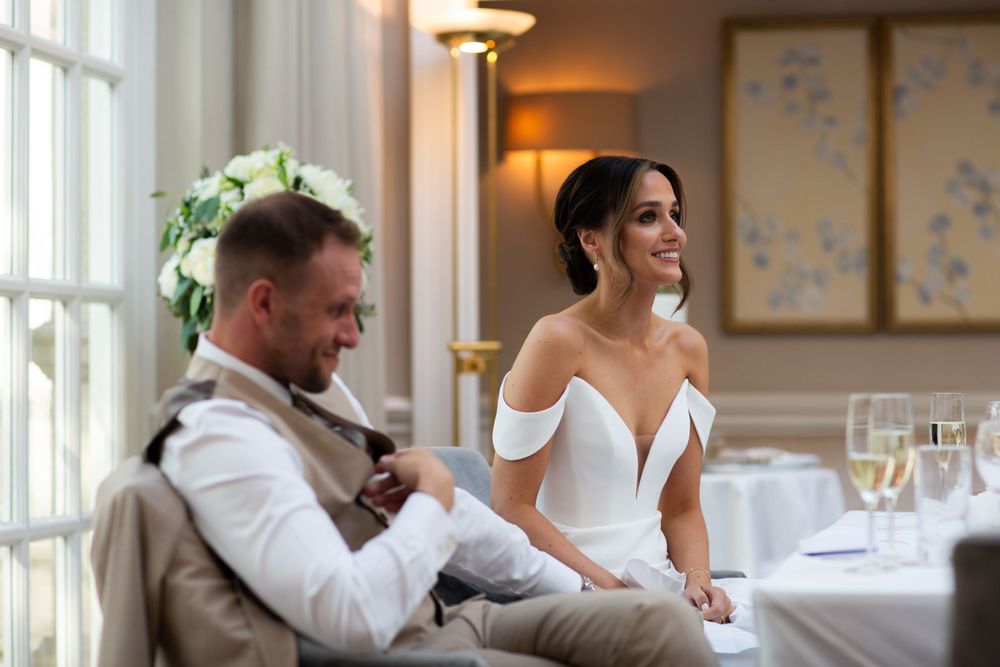 A bride and groom sit together at their elegant wedding reception in a warmly lit room with floral wall art.