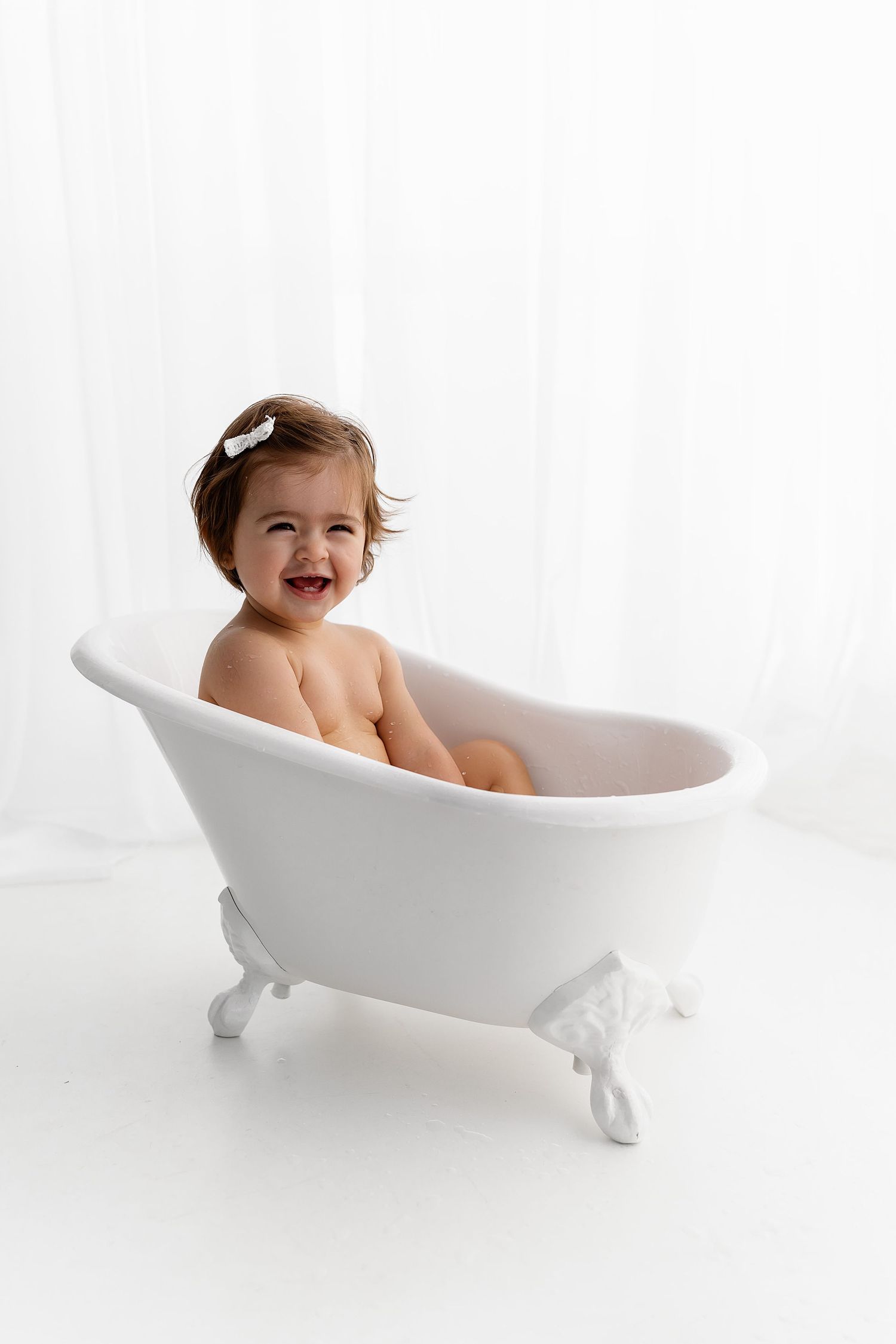 Toddler laughs joyfully while sitting in vintage-style white bathtub.