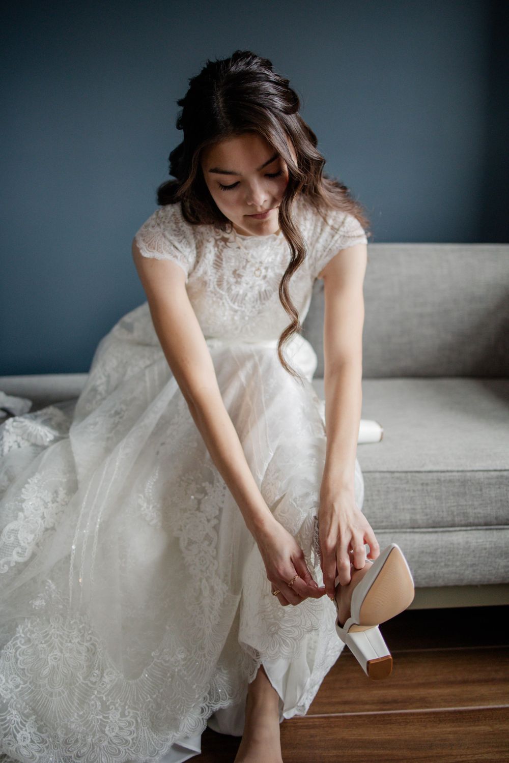 Bride in lace cap sleeve dress sits on grey couch while putting on white heels.