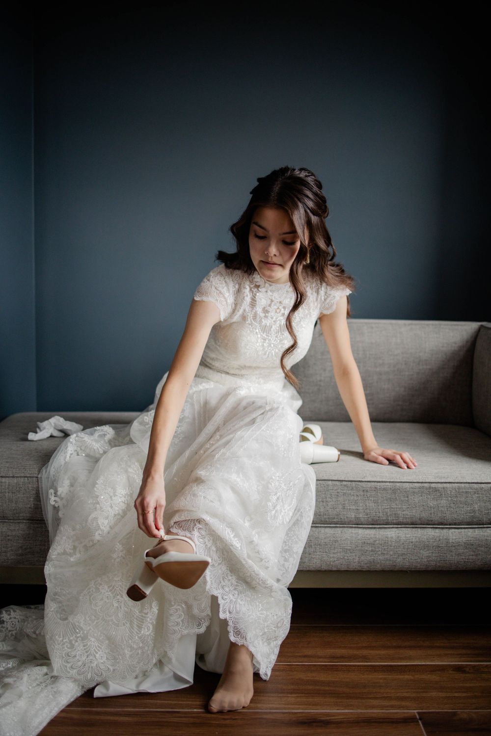 Elegant bride in white lace dress sits thoughtfully on grey sofa in moody lighting.