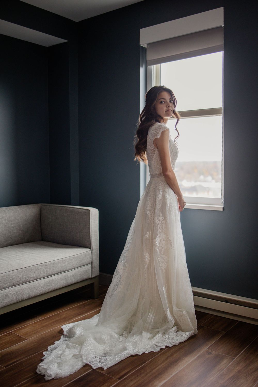 Bride in flowing lace gown poses by window in room with grey walls and hardwood floors.