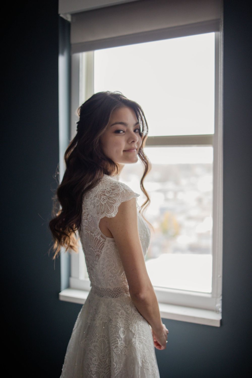 Backlit bride stands by window in cap sleeve lace wedding gown with flowing train.