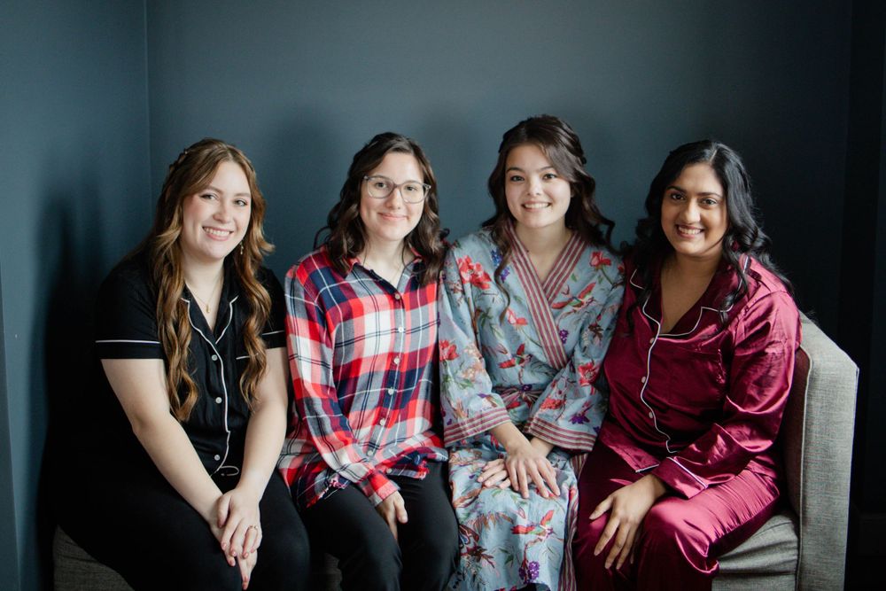 Four women in casual wear and pajamas smile while sitting together on couch.