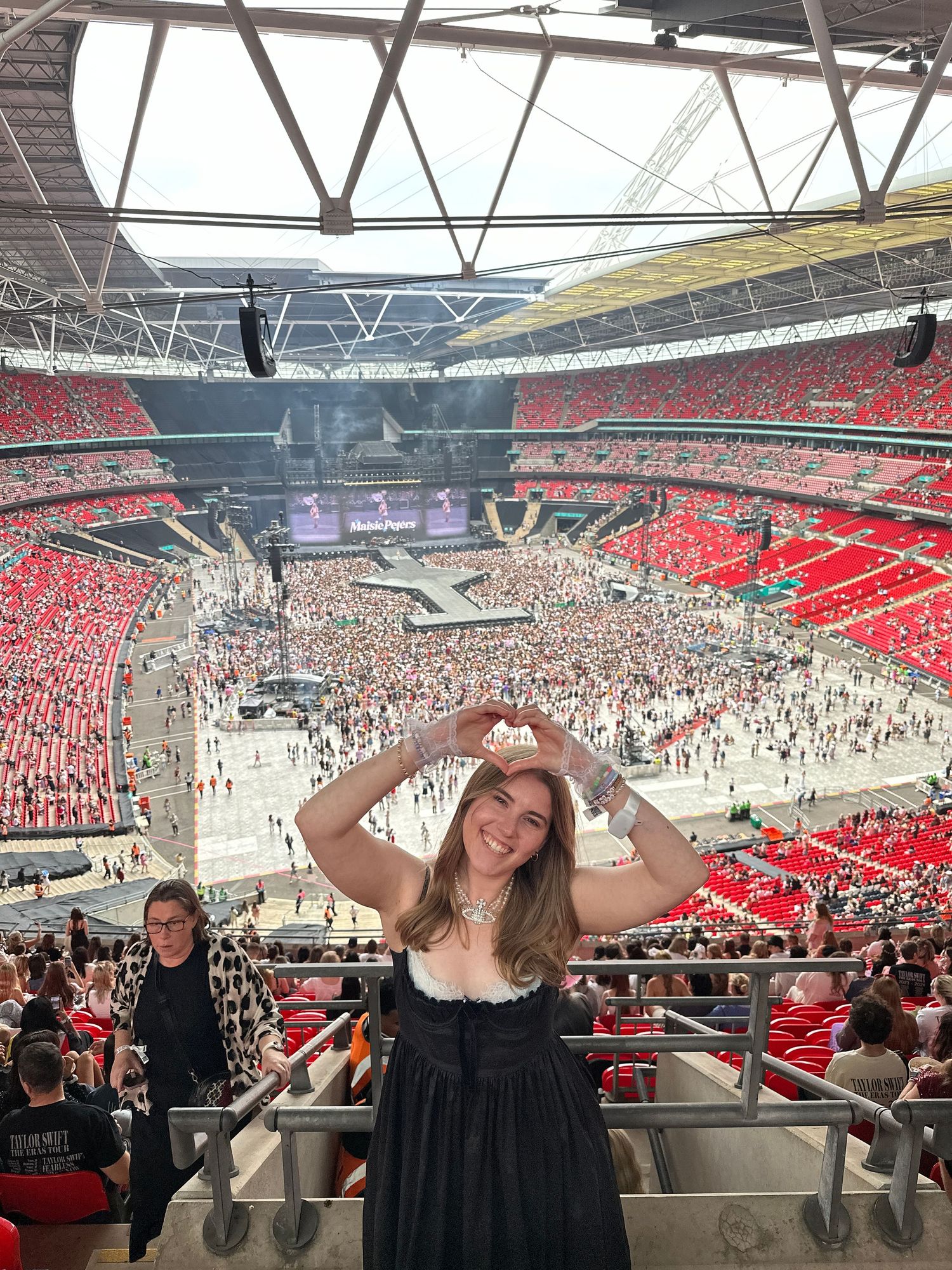 Concert attendee poses at Wembley Stadium with a heart gesture amid a massive crowd.