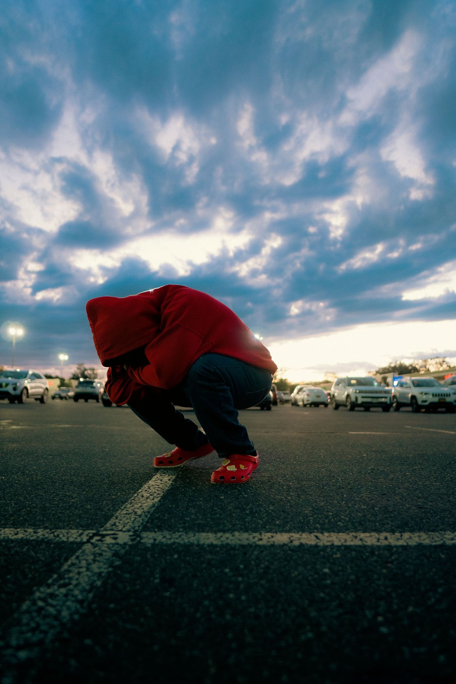 Figure in a red hoodie crouching in a parking lot at sunset with dramatic cloudy sky above.