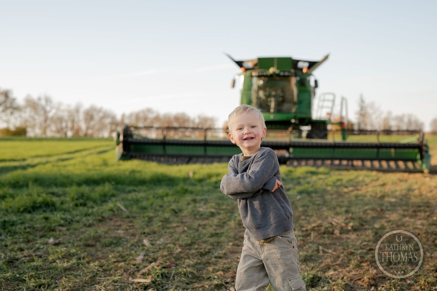 A joyful child poses with crossed arms in front of a large green combine harvester.