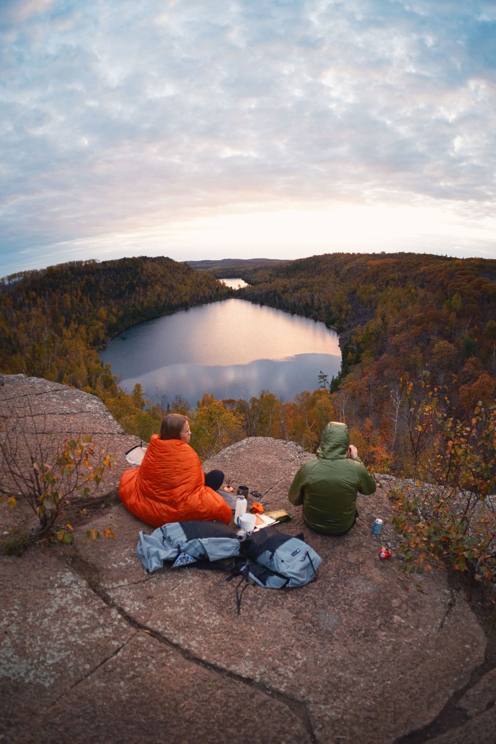 Hikers sitting on a rocky overlook enjoying scenic views of a lake surrounded by autumn forest at sunset.