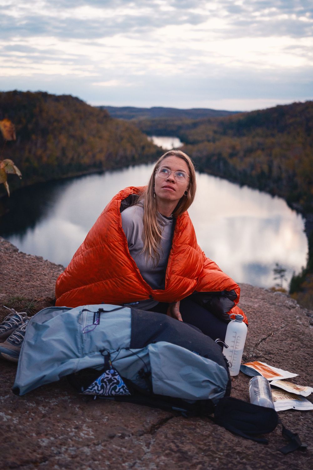 Hiker relaxing with camping gear on rocky overlook above scenic lake at sunset in autumn mountains.