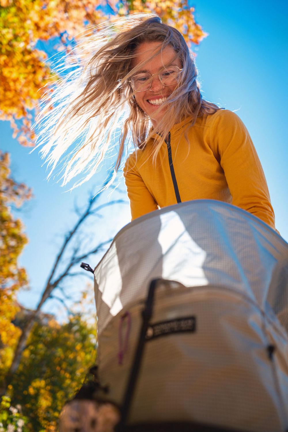 Person in yellow sweater smiling and playing with autumn leaves against bright blue sky on sunny fall day.