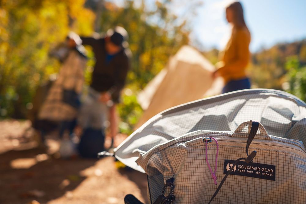 Close-up of a camping backpack with people setting up a tent in the blurred background on a sunny day.