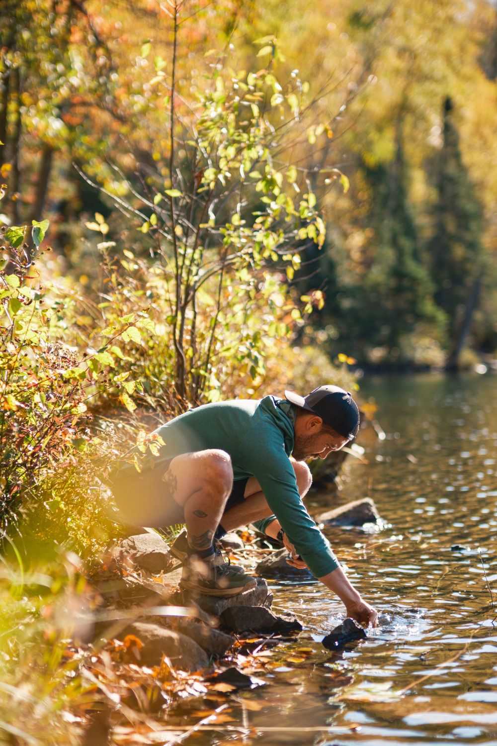 Person in teal sweater reaches into a tranquil lake surrounded by autumn foliage on a sunny day.