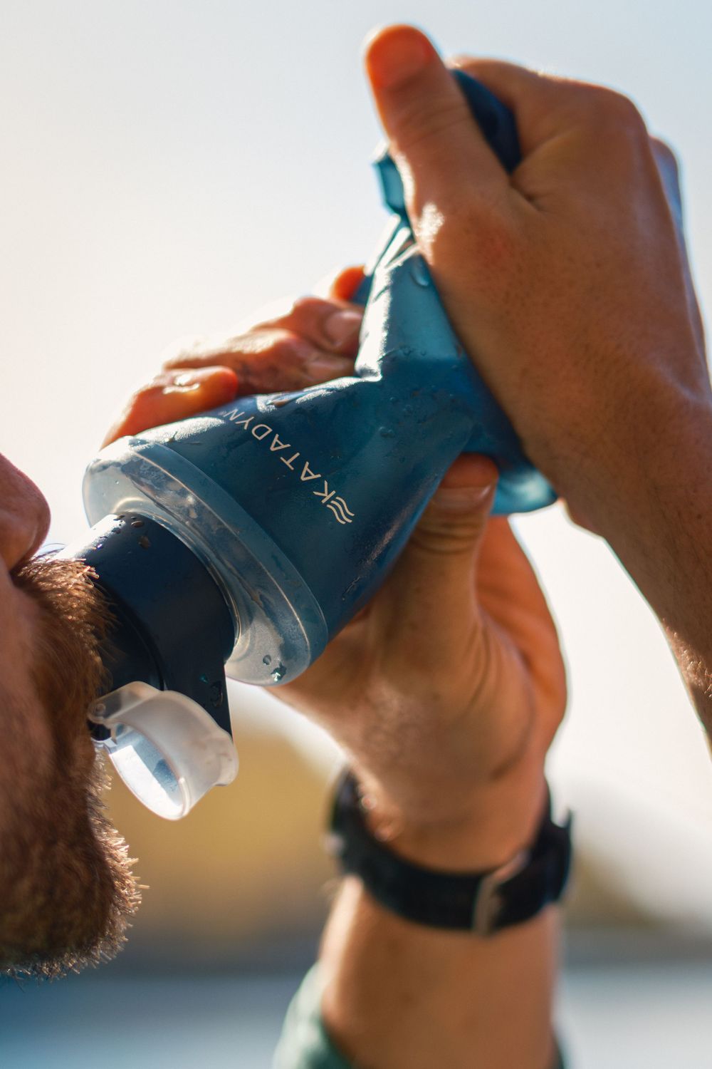 Close-up of hands holding and drinking from a blue water bottle against bright sky.