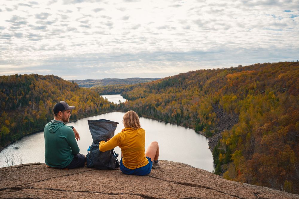 Two people sit on cliff edge overlooking winding river valley with autumn foliage and cloudy sky.
