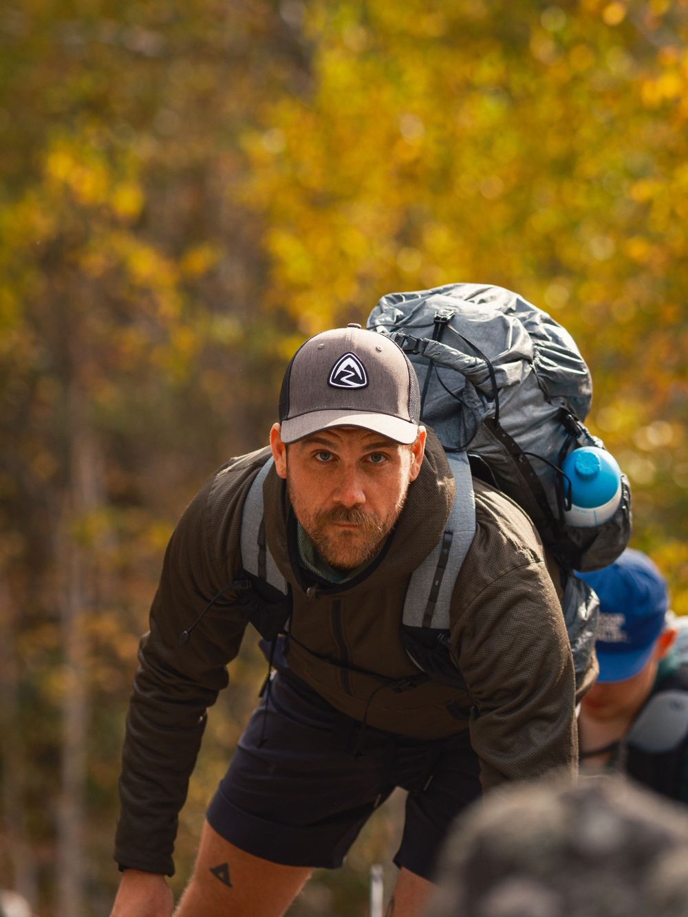 Hiker wearing a baseball cap and backpack climbs uphill against backdrop of golden autumn foliage.