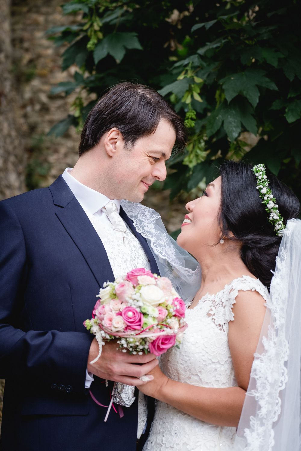 A bride and groom share an intimate moment while holding a pink rose bouquet against a leafy backdrop.