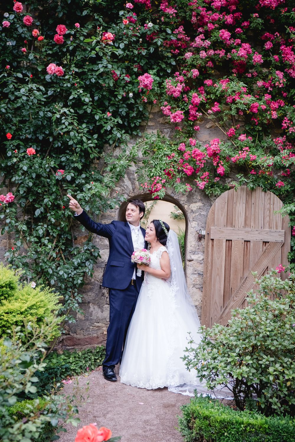 A couple poses beneath an archway covered in climbing pink roses and a rustic wooden gate.