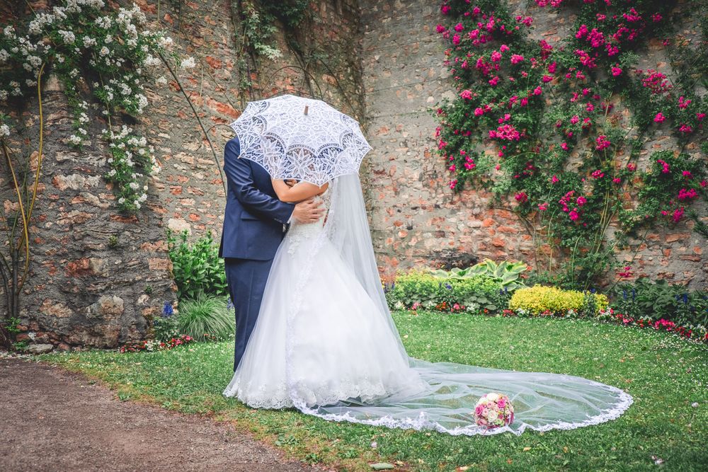 A wedding couple embraces against a stone wall covered with climbing roses in a garden setting.